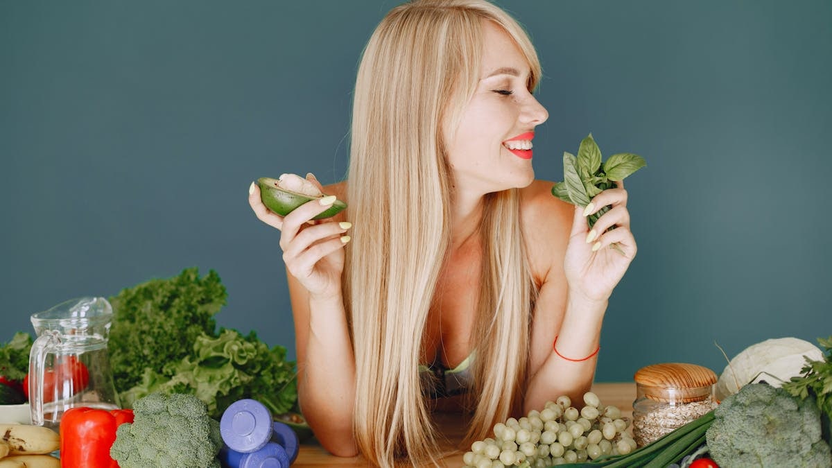 A smiling woman holding avocado and basil, surrounded by fresh vegetables and fruits for healthy eating.